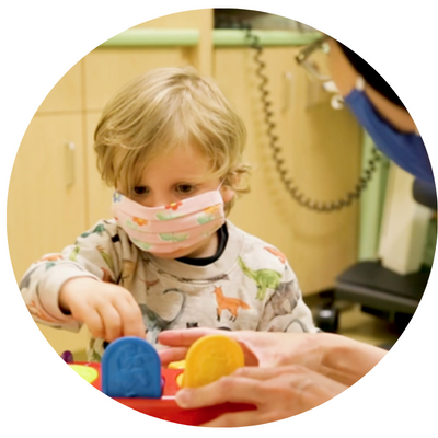 A child is wearing a mask and playing with a toy while an adult looks on. 